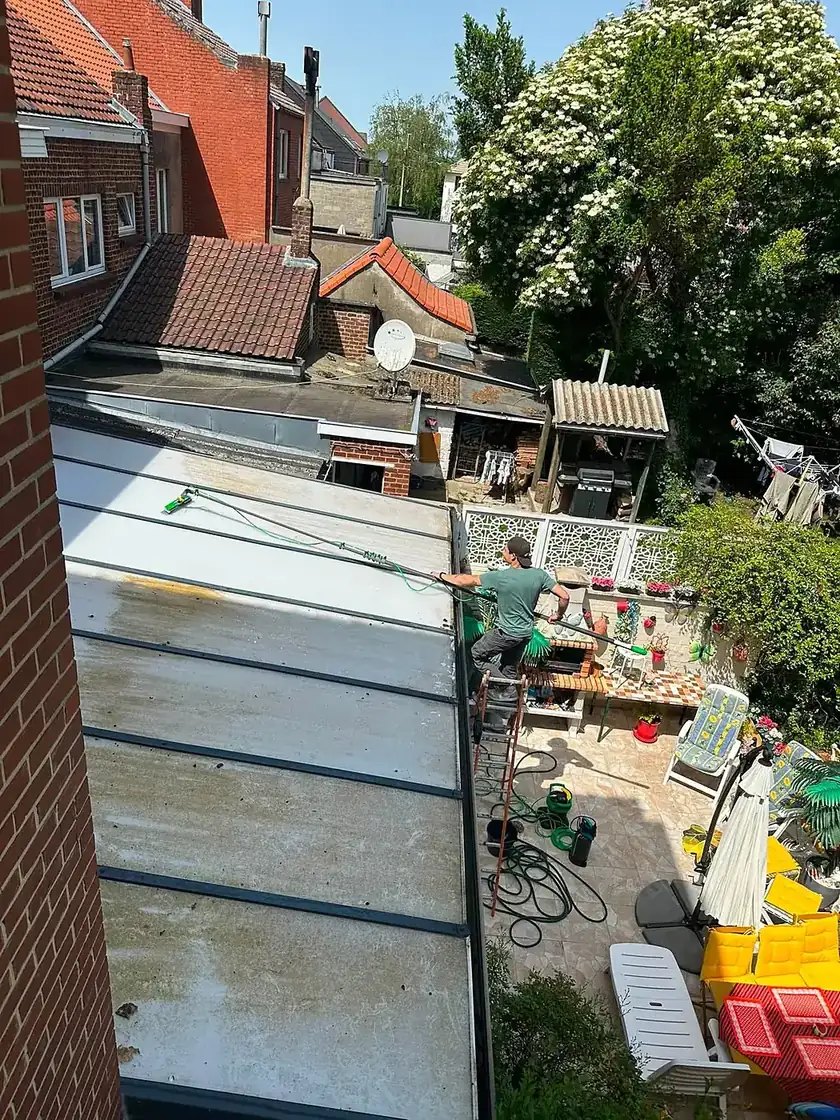 Nettoyage d'une toiture de véranda à la perche télescopique, depuis une échelle posée au bord du toit.